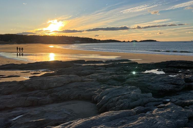 A scenic photo of the Gulf of Maine shoreline showing two people walking into a sunset.