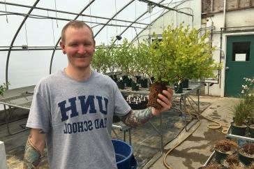 Male student in greenhouse holds a plant. His t-shirt says 主要研究 研究生院.