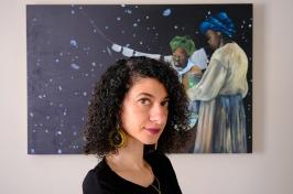 Woman with dark curly hair looks at the camera, painting of Black astronomers behind her