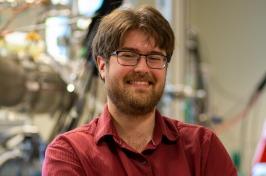 主要研究 PhD candidate David Ruth smiles at the camera in front of physics equipment