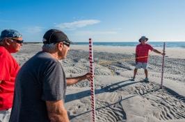 Three elderly men stand on a beach holding measuring sticks to determine height of the sand.