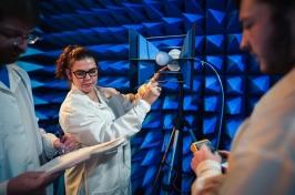 Three people wearing white lab coats 和 holding space testing equipment st和 in a room full of blue foam cones.